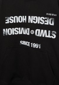 Črna tkanina z belim, obrabljenim besedilom "STWD © DMISION DESIGN HOUSE SINCE 1991" in "01/02 STAFF," delno obrnjena na glavo.