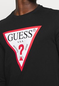 Schwarzes Langarmshirt mit einem großen weißen und roten dreieckigen GUESS-Logo, das ein rotes Fragezeichen und den Schriftzug "WASHED JEANS" auf der Brust zeigt.