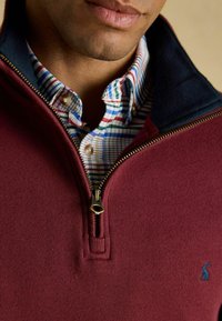 Bordeaux ritsweater met een geribbelde textuur, marineblauwe kraag, gouden rits, over een meerkleurig geruit overhemd met blauwe, groene en rode accenten.