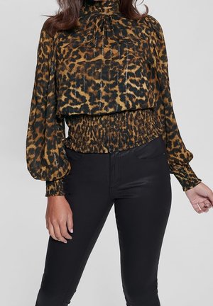 Kvinde iført en leopardprint langærmet bluse med smockdetaljer i talje og manchetter, kombineret med sorte tætsiddende bukser mod en ensfarvet baggrund.