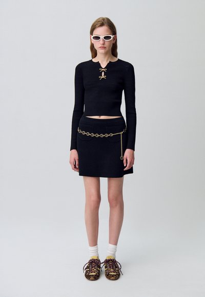 Top corto de punto negro con detalle de lazo, combinado con una falda mini negra que presenta un cinturón de cadena dorada. El calzado incluye zapatillas con estampado y calcetines blancos.