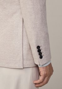 Blejzer bej deschis dintr-un material texturat. Prezintă un singur slit, patru nasturi negri la manșetă și o căptușeală contrastantă a mânecii dedesubt.