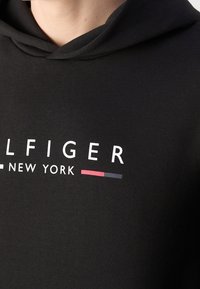 Svart huvtröja med en framficka, dragsko på huvan och en vit och färgglad logotyp som säger "TOMMY HILFIGER - NEW YORK." Slät tygkvalitet.