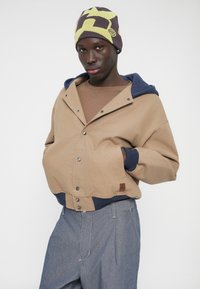 Béžová mikina s kapucňou s tmavomodrými prvkami, zapínaním na cvoky a náplasťou. Nosená cez hnedú košeľu, v kombinácii s modrými rifľovými nohavicami a vzorovanou čiapkou.