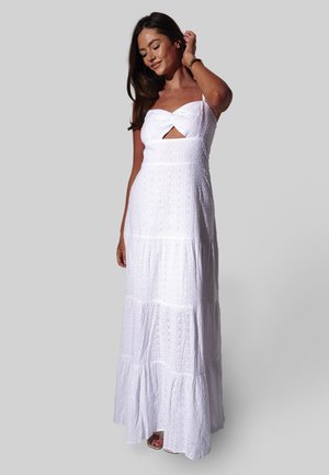 Vrouw met lang, donker haar, gekleed in een witte mouwloze maxijurk met ajourpatroon en een uitsnijding aan de voorkant, staande tegen een effen grijze achtergrond.