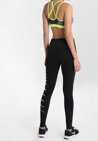 Leggings nere con accenti giallo neon e design aderente, abbinate a un reggiseno sportivo grigio con spalline incrociate sulla schiena.