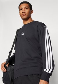 Sweatshirt preto com mangas longas e riscas brancas nas laterais, apresentando um pequeno logótipo branco da Adidas no peito. Textura de tecido suave.