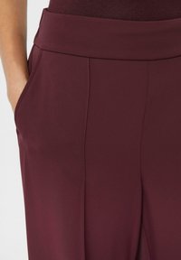 Bordeauxfarbene Hose mit einem breiten Bund, ausgestattet mit seitlichen Taschen und einer glatten, weichen Stoffoberfläche. Maßgeschneidertes Design mit klaren Linien.
