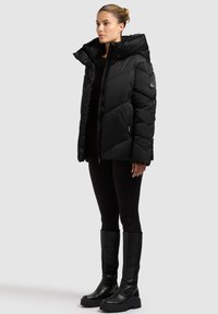 Schwarze Steppjacke mit hohem Kragen, Reißverschluss und gestepptem Muster. Trägt sich über einem schwarzen Oberteil und wird mit kniehohen schwarzen Stiefeln kombiniert.