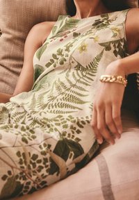 Femme allongée sur une chaise, portant une robe sans manches à imprimé floral et feuilles vertes, avec un gros bracelet en or à son poignet.