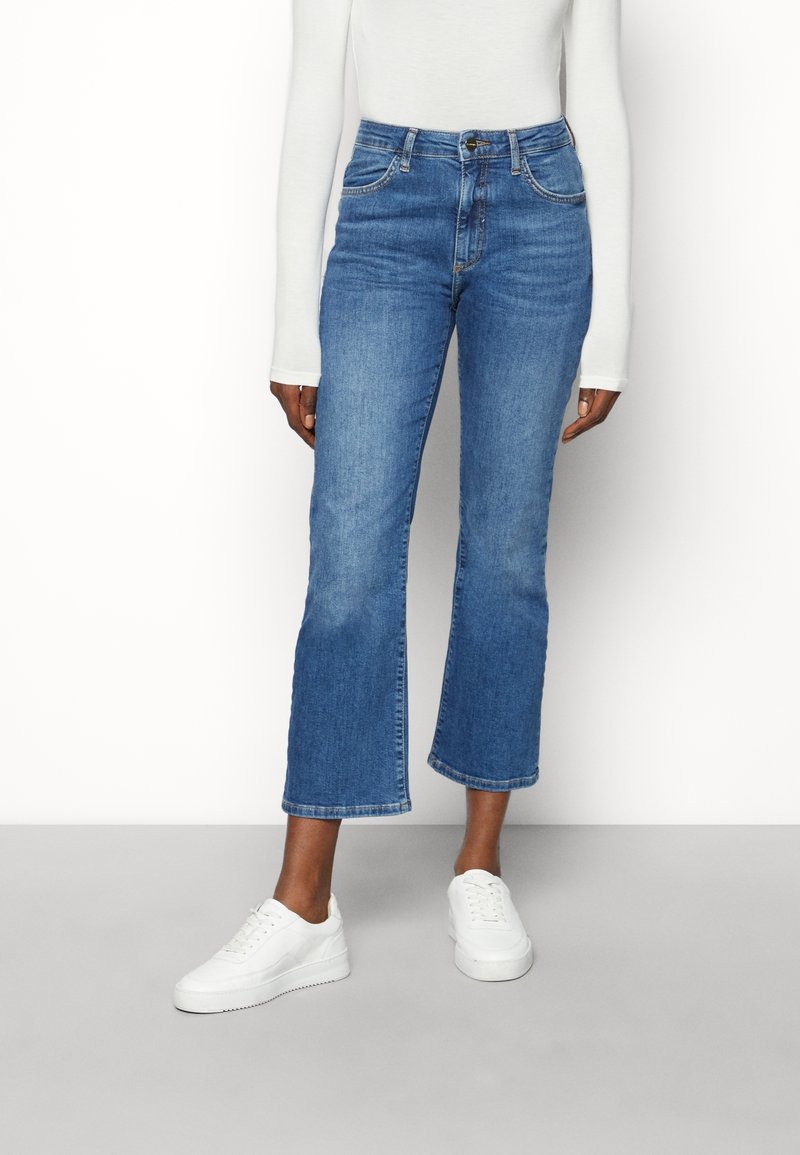 Blå denim jeans med vida ben och hög midja, med framfickor och rå kant. Matchas med vita sneakers för en avslappnad look.