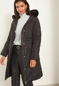 Černá péřová bunda s kapucí lemovanou kožešinou, prošívaný design, přední zip a zlaté kovové doplňky, nošená přes šedý svetr a černé kalhoty.