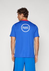 Blå atletisk T-shirt med et rundt hvidt logo med "PARIS" på ryggen. Rød detalje ved halsudskæringen, korte ærmer og letvægtsstof.