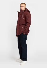 Bordeauxkleurige jas met een capuchon, voorzakken, zilveren drukknoopsluitingen en een trekkoord. Gecombineerd met marineblauwe cargobroeken en witte sneakers.