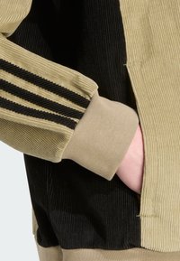 Corduroy jacket featuring black and beige panels, with ribbed texture and three black stripes on the sleeve. Adjustable cuffs included.