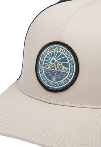 Beige kasket med en rund skygge. Har et broderet cirkulært patch med bjerge og sol, kantet i sort. Teksten lyder "Billabong Adventure Division."
