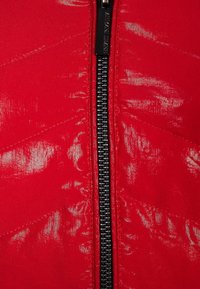 Röd vadderad jacka med en svart dragkedja och ett chevron-mönster, med en glansig, texturerad yta.