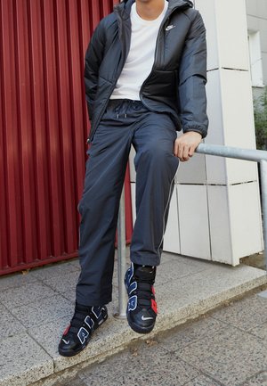 Personne vêtue d’une veste noire Nike, d’une chemise blanche, d’un pantalon foncé et de baskets noires Nike, assise sur une rambarde métallique à côté d’un bâtiment rouge et blanc.