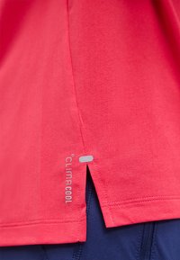Ourlet rouge de chemise athlétique avec fente latérale et logo CLIMACOOL réfléchissant sur tissu de shorts ou leggings bleu marine.