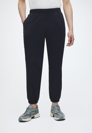Personne portant un pantalon jogger noir avec des poignets élastiques, une chemise blanche et des baskets de sport grises, debout avec une main dans la poche.