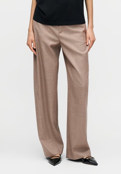 Pantalones beige de pierna ancha hechos de tela texturizada, con un corte entallado y un patrón sutil, combinados con zapatos negros de punta afilada.