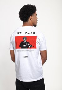 Hvid bomulds t-shirt med et rødt grafisk design på ryggen, som indeholder tekst på japansk og engelsk, samt en sort illustration af en mand, der holder et våben.