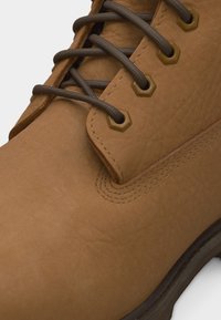 Κοντινό πλάνο από μια μπεζ δερμάτινη μπότα με σκούρα καφέ κορδόνια περασμένα μέσα από εξαγωνικές οπές και ορατές λεπτομέρειες ραφής.