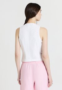 Weißes ärmelloses Oberteil mit Lochmuster, kurzes Design und strukturiertem Stoff. Kombiniert mit rosa elastischen Hosen, von hinten betrachtet.