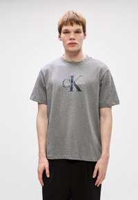 Γκρι βαμβακερό μπλουζάκι με κοντά μανίκ�ια, που φέρει το λογότυπο "Calvin Klein" και το "ck" σε αντίθετο σκούρο γράμμα στο μπροστινό μέρος.