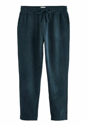 Femme portant une chemise en velours côtelé bleu marine boutonnée et un pantalon assorti avec un crop top blanc et des chaussures beiges à enfiler à l’intérieur.