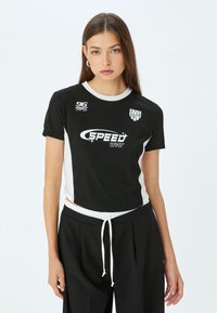 Sort cropped t-shirt med hvide sidepaneler. Har logo tekst "SPEED" og numre trykt på forsiden. Korte ærmer, rund halsudskæring.