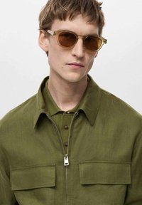Olive green, textured shirt with two chest pockets and a zippered collar, worn with transparent-framed sunglasses and a soft beige undershirt.
