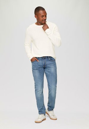 Chemise blanche à manches longues, jeans en denim bleu clair et baskets blanches avec des accents beiges. La tenue présente une coupe décontractée et un style casual.