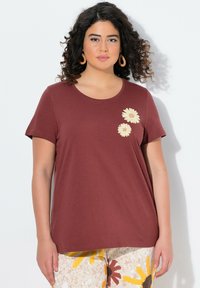 Camiseta de manga corta color burdeos que presenta tres margaritas bordadas de colores claros en el lado izquierdo, confeccionada con un material suave y liso.