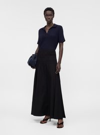 Femme portant un haut en maille manches courtes bleu marine, une longue jupe noire, des sandales à talons noires et tenant un sac à main bleu marine sur un fond uni.