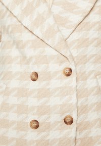 Beige kappa med houndstooth-mönster och stora slag, dubbelknäppt design och fyra runda, beigea knappar. Mjuk, strukturerad tyg.