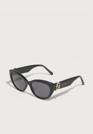 Juodos kačių akių saulės akiniai su tamsiais lęšiais ir auksinės spalvos Marc Jacobs logotipu plačiose šoninėse dalyse, parodyti ant vientisos šviesios fono.