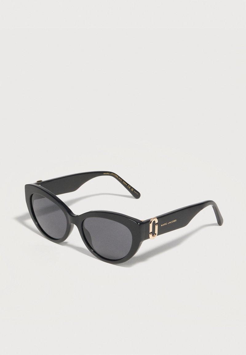 Μαύρα γυαλιά ηλίου σε σχήμα γάτας με σκούρους φακούς και χρυσό λογότυπο Marc Jacobs σε πλατιά πλαϊνά, παρουσιασμένα σε απλό ανοιχτό φόντο.