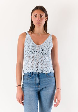 Donna con top senza maniche in maglia blu e jeans blu a vita alta, in piedi davanti a uno sfondo bianco uniforme, rivolta in avanti con espressione neutra.