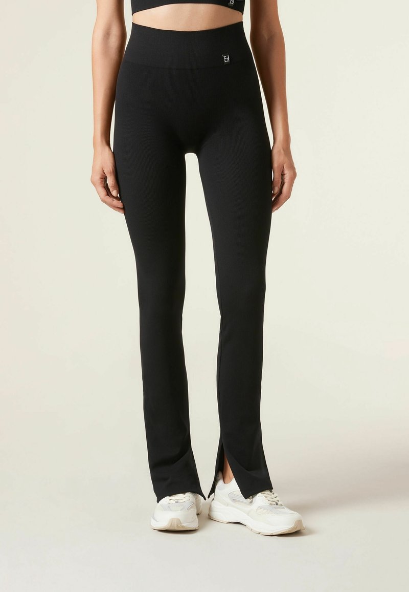 Femme portant des leggings noirs taille haute avec une fente à l'avant de l'ourlet et des baskets blanches, debout devant un fond uni.