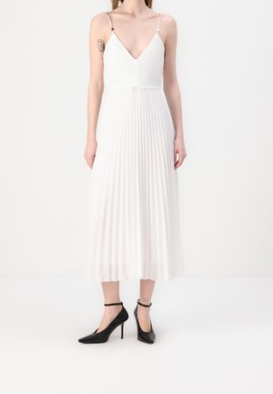 Mujer con un vestido midi plisado blanco con tirantes finos y tacones altos negros con correa en el tobillo, de pie frente a un fondo liso.