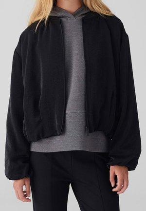Femme portant une veste noire zippée sur un sweat à capuche gris et un pantalon noir, debout devant un fond clair uni.