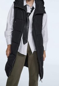 Gilet imbottito nero con cappuccio e chiusura con zip, caratterizzato da un design trapuntato, indossato sopra una maglia a righe e pantaloni verde oliva.