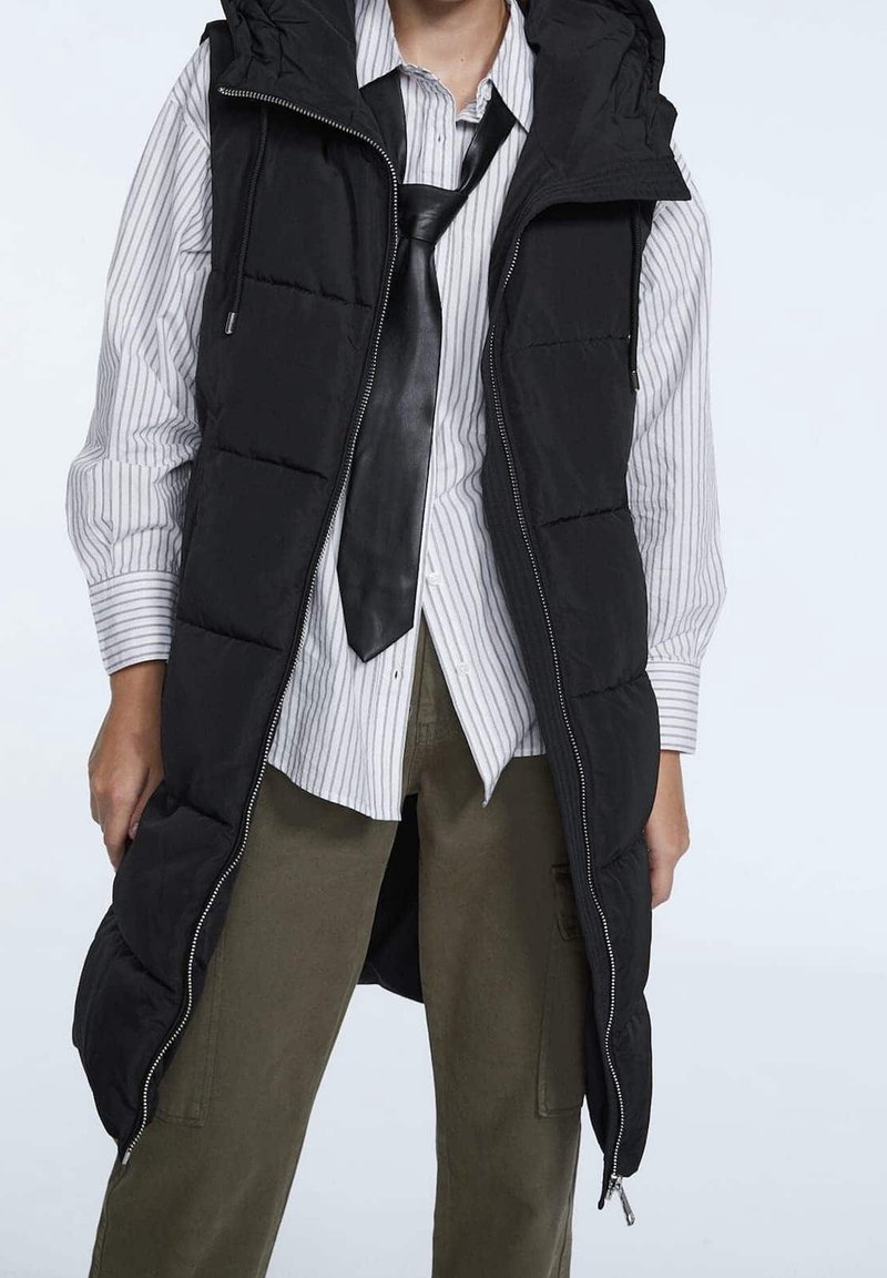 Gilet imbottito nero con cappuccio e chiusura con zip, caratterizzato da un design trapuntato, indossato sopra una maglia a righe e pantaloni verde oliva.