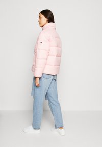 Rosa boblejakke med horisontalt quiltet design, høy krage og logo på ermet, kombinert med lyseblå jeans og hvite joggesko.
