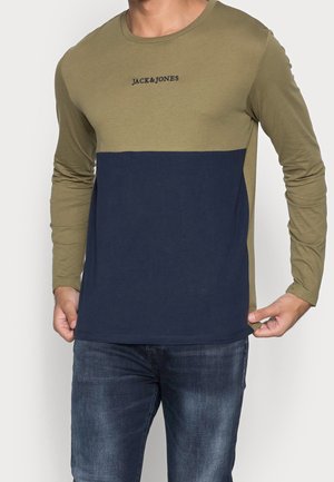 Mężczyzna ubrany w koszulę z długim rękawem w kolorowe bloki: oliwkowa góra, granatowa dolna część i logo "JACK & JONES", zestawioną z niebieskimi dżinsami.