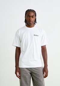Hvit bomull t-skjorte med korte ermer har en liten svart "Dickies"-logo på øvre venstre side. Kombinert med grå bukser.