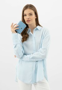 Camicia a righe azzurra con chiusura a pulsante e maniche arrotolate, abbinata a un piccolo portafoglio blu testurizzato tenuto nella mano destra.