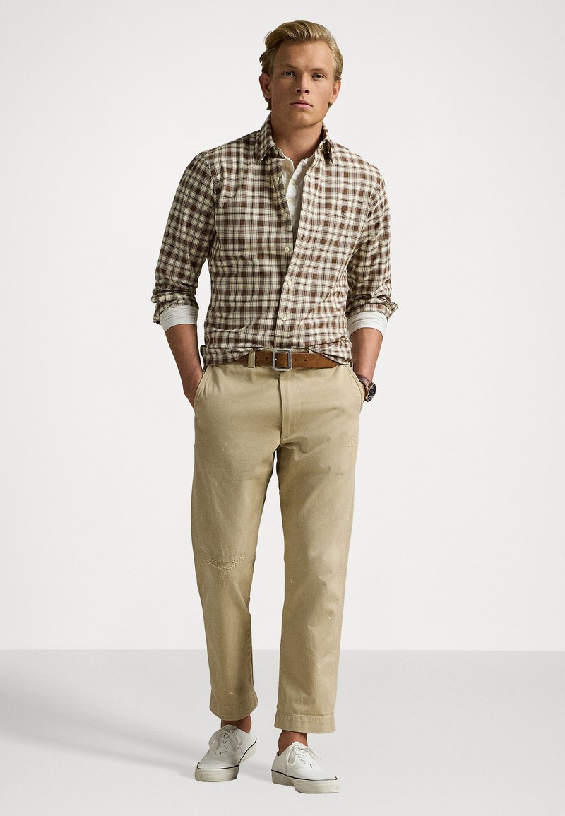Camisa de cuadros marrón y blanco, pantalones beige con detalles desgastados, cinturón marrón y zapatillas blancas. Ajuste casual, diseño de botones.
