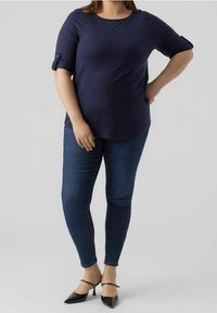 Marineblaues, kurzärmliges Oberteil mit umgeschlagenen Manschetten, kombiniert mit dunkelblauen Skinny-Jeans und schwarzen spitzen High Heels. Glatte Stofftextur.